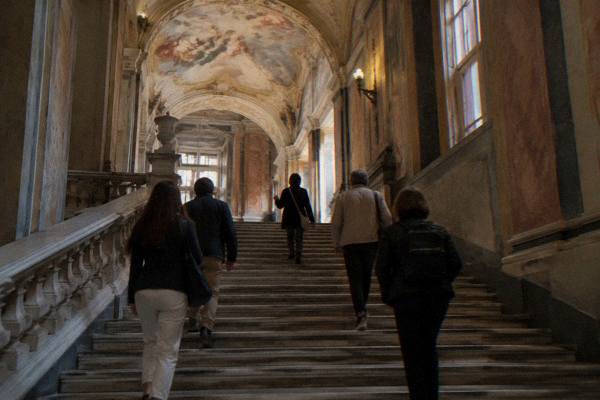 A small group climbing a wide marble staircase of a grand palace behind a guide, seen from behind, painted vaulted ceiling above, Palazzi dei Rolli, Genoa